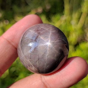 May include: A polished, dark gray star sapphire sphere with a six-pointed star visible in the center. The sphere is smooth and reflective, with a slight sheen.
