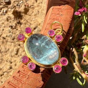 May include: A gold pendant featuring a large oval aquamarine gemstone, encircled by smaller pink gemstones. The pendant has a gold bail for attaching to a chain.