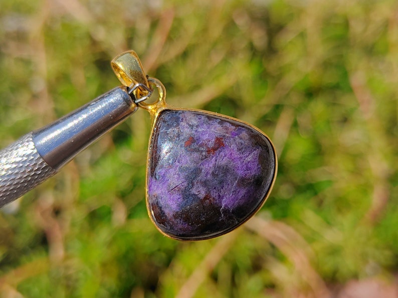 Pode incluir: Um pingente em forma de l&aacute;grima com uma pedra preciosa roxa e preta engastada em uma moldura de metal dourado.