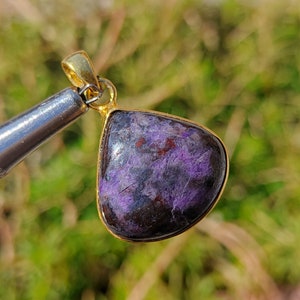 Pode incluir: Um pingente em forma de l&aacute;grima com uma pedra preciosa roxa e preta engastada em uma moldura de metal dourado.