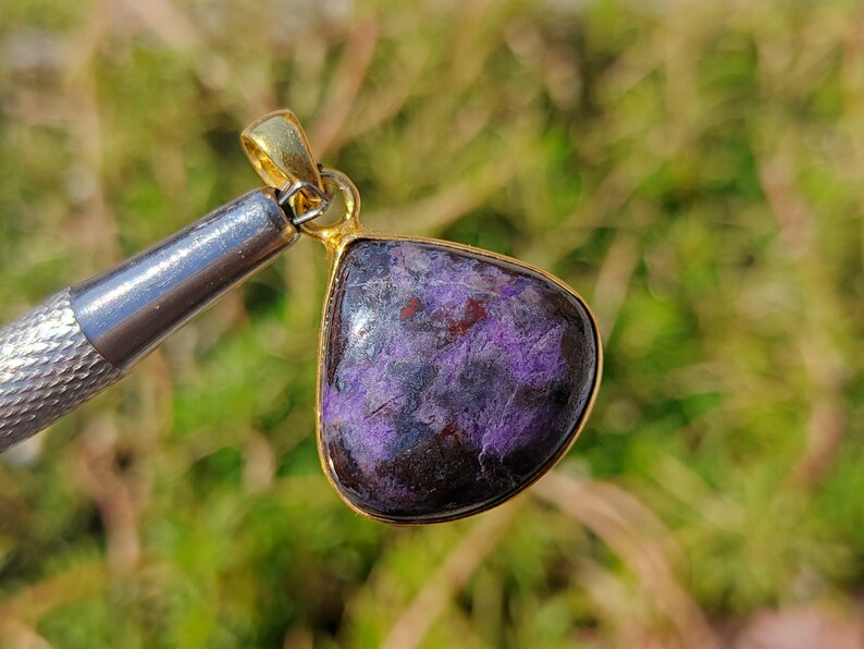 Pode incluir: Um pingente em forma de l&aacute;grima com uma pedra preciosa roxa e preta engastada em uma moldura de metal dourado.