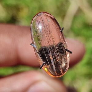 May include: A translucent, oval-shaped, pink gemstone with visible internal lines. The gemstone is held in place by four metal prongs.
