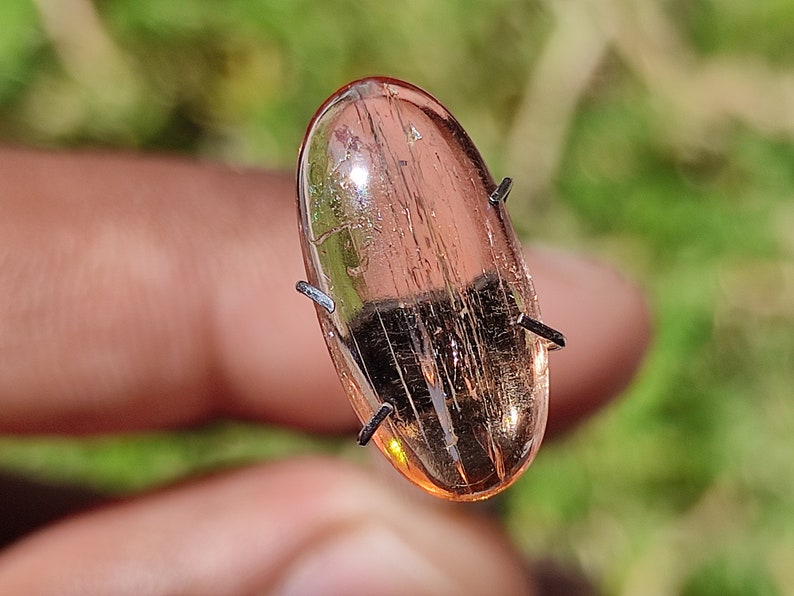 May include: A translucent, oval-shaped, pink gemstone with visible internal lines. The gemstone is held in place by four metal prongs.