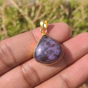 Pode incluir: Um pingente em forma de l&aacute;grima com uma pedra preciosa roxa e preta engastada em uma moldura de metal dourado.