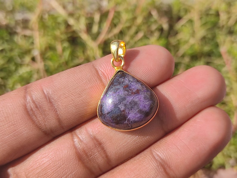 Pode incluir: Um pingente em forma de l&aacute;grima com uma pedra preciosa roxa e preta engastada em uma moldura de metal dourado.