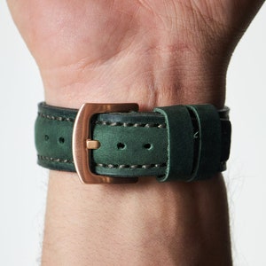May include: A green leather watch strap with a rose gold buckle. The strap has white stitching and is shown on a person's wrist.