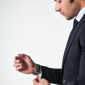 May include: A man in a black suit adjusts the cuff of his sleeve, revealing a green leather wristwatch.