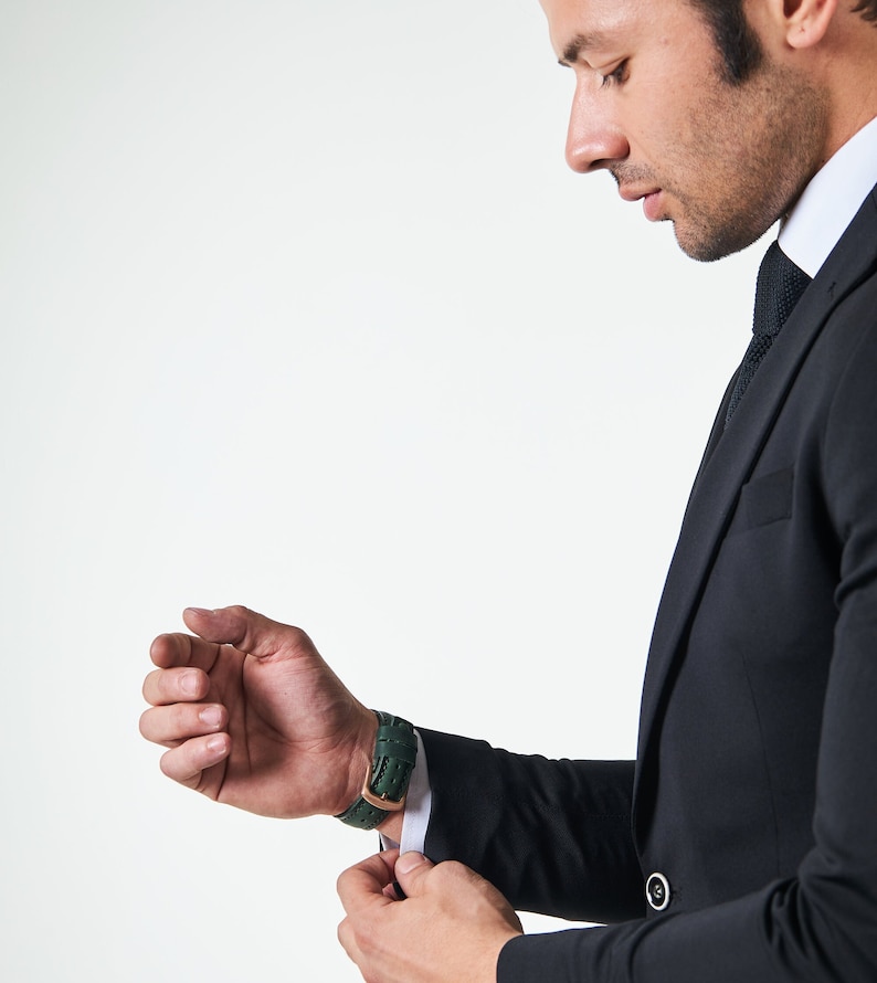 May include: A man in a black suit adjusts the cuff of his sleeve, revealing a green leather wristwatch.