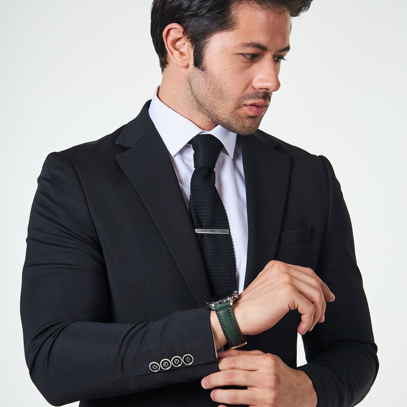 May include: A man wearing a black suit jacket with a white shirt and a dark green knitted tie. He is wearing a silver tie bar and a green leather watch band.