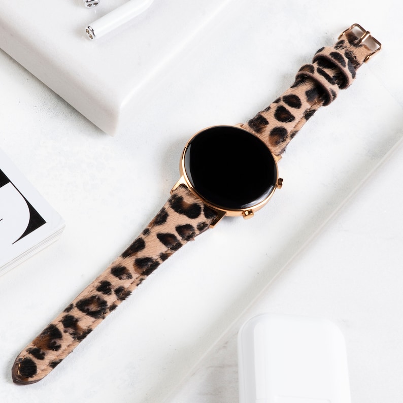 Puede incluir: Un reloj inteligente dorado con una esfera negra y una correa de estampado de leopardo.