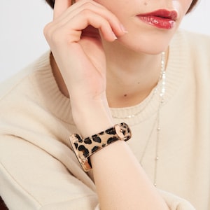 Puede incluir: Un smartwatch de oro rosa con una banda de estampado de leopardo. La banda est&aacute; hecha de un material suave y peludo.