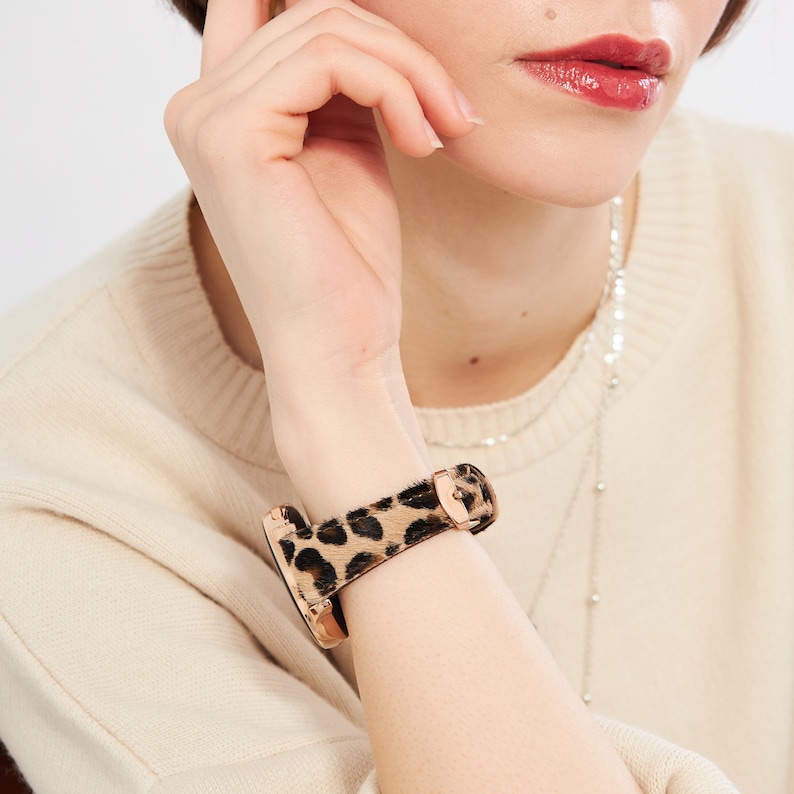 Puede incluir: Un smartwatch de oro rosa con una banda de estampado de leopardo. La banda est&aacute; hecha de un material suave y peludo.
