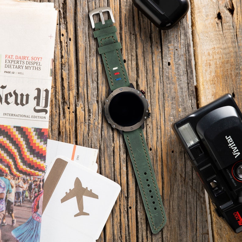 May include: A green leather watch band with red, white, and blue accents. The watch band is attached to a black and silver smartwatch with a round face.