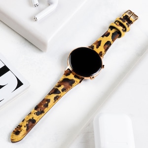 Puede incluir: Un reloj inteligente dorado con una esfera negra y una banda de estampado de leopardo. La banda est&aacute; hecha de un material suave y texturizado.