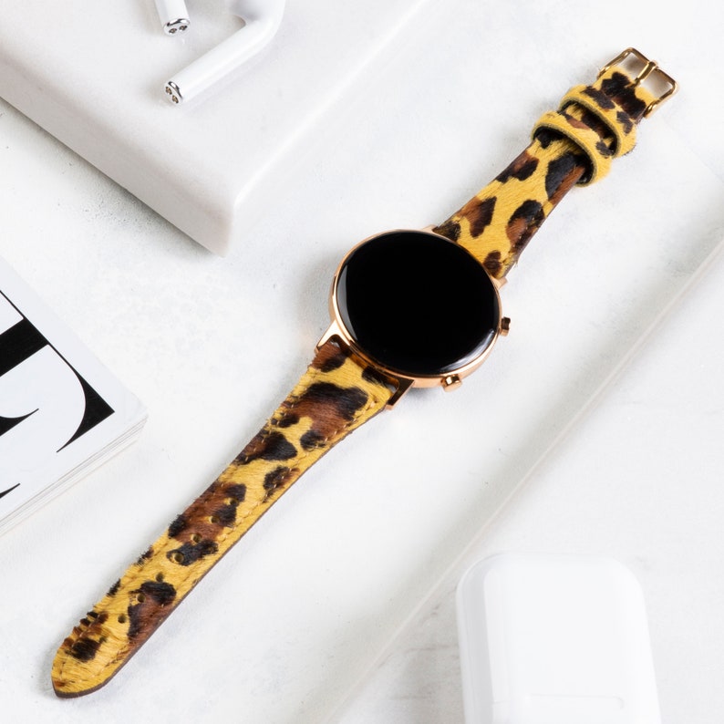 Puede incluir: Un reloj inteligente dorado con una esfera negra y una banda de estampado de leopardo. La banda est&aacute; hecha de un material suave y texturizado.