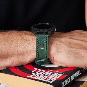 May include: A green leather watch band with white, red, and blue stitching. The band is attached to a black smartwatch with a round face.