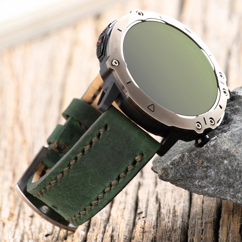 May include: A green leather watch band with stitching detail. The band is attached to a silver smartwatch with a black bezel and a round, dark green screen.