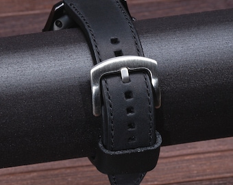 Peut inclure: Un bracelet de montre en cuir noir avec une boucle argentée. Le bracelet est enroulé autour d'un rouleau de tissu noir.