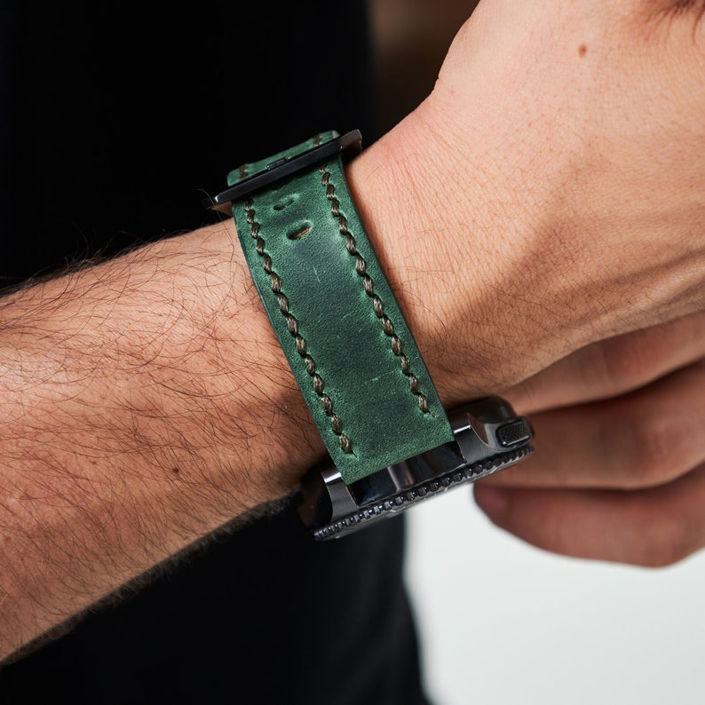 May include: A dark green leather watch band with white stitching. The band is attached to a black smartwatch.