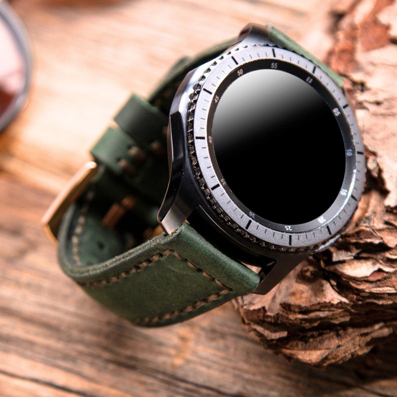 May include: A green leather watch band with a black smartwatch with a round face and a silver bezel. The watch face has numbers from 40 to 60 around the edge.