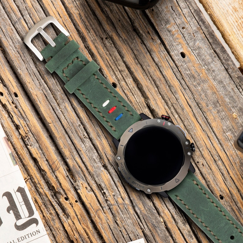 May include: A green leather watch band with white stitching and red, white, and blue accents. The band is attached to a black smartwatch with a round face and a silver bezel. The watch face is black and has a white digital display. The watch is on a wooden surface.