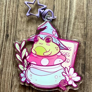 Può includere: Un portachiavi in acrilico trasparente con un ranocchio dei cartoni animati con un cappello da strega, seduto su un fungo rosa. Il portachiavi ha una chiusura a forma di stella viola e include un cartello con la scritta "Froggies".