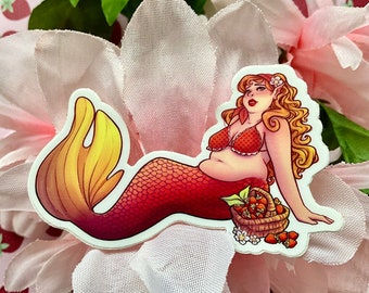 May include: A sticker of a mermaid with red hair and a red and orange tail, wearing a red bikini top. She is reclining next to a basket of strawberries. The sticker is placed on a pink flower with green leaves.