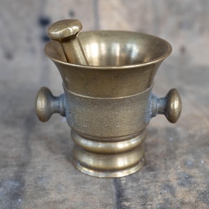 May include: A vintage brass mortar and pestle set. The mortar has a flared rim, two side handles, and a rounded base. The pestle rests inside the mortar. The set has a tarnished, antique finish.