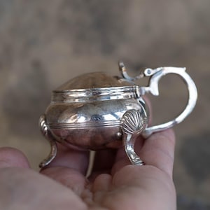 Peut inclure: Une salière en argent avec un couvercle et trois pieds. La salière a un corps arrondi et une poignée décorative.