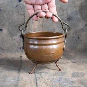 Peut inclure: Un petit chaudron en cuivre antique avec un corps arrondi et trois courtes pattes. Il est doté d'une poignée incurvée en métal plus foncé, fixée sur les côtés. Le chaudron a une surface polie et réfléchissante, montrant des signes d'âge et d'utilisation.
