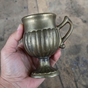 Puede incluir: Una pequeña taza de latón antiguo con asa y cuerpo acanalado. La taza tiene una base de pedestal y un borde ligeramente ensanchado. El latón tiene un aspecto deslustrado y envejecido, lo que sugiere que es vintage.