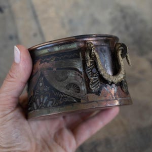 Peut inclure: Une petite casserole ovale en cuivre avec des motifs gravés complexes. La casserole a deux anses en laiton avec des lanières de cuir. La casserole est de couleur cuivre foncé avec des touches d'argent et d'or.