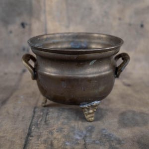 Könnte beinhalten: Ein kleiner antiker Topf aus Messing mit drei Beinen und zwei Griffen. Der Topf hat eine abgenutzte, gealterte Oberfläche und ist perfekt für dekorative Zwecke.