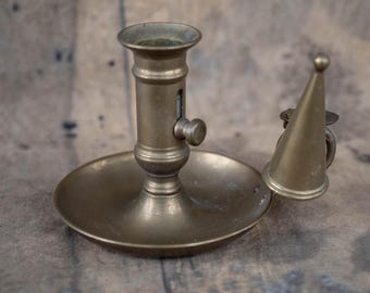 May include: Antique brass candlestick with a round base and a cylindrical stem. A conical snuffer sits beside the candlestick. The metal has a warm, aged patina. The candlestick is designed to hold a single candle.