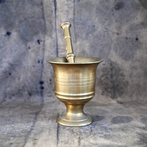 Peut inclure: Un mortier et un pilon en laiton. Le mortier a un bord évasé et une base à pied. L'extérieur est orné de bandes horizontales. Le pilon est cylindrique avec une poignée arrondie. L'ensemble est sur un fond gris moucheté.