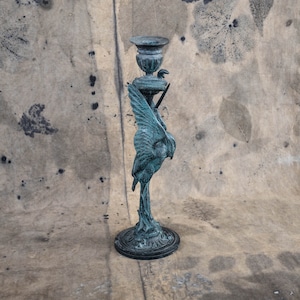May include: A bronze-colored candlestick holder with a bird design. The candlestick has a round base, a slender stem, and a cup-shaped top. The bird is depicted with outstretched wings, adding a decorative element to the piece.