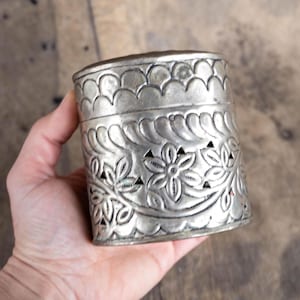 May include: A cylindrical silver-toned metal container featuring an embossed pattern of scalloped edges at the top and bottom, with a central band displaying intricate floral motifs and cut-out details throughout the surface of the vessel.