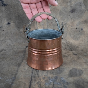 Könnte beinhalten: Ein kleiner Kupferkübel mit Henkel. Der Kübel hat ein abgenutztes, altes Aussehen und ist aus gehämmertem Kupfer gefertigt.