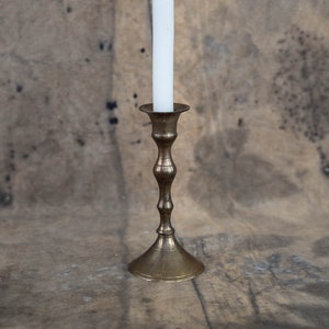 Könnte beinhalten: Ein Messing-Kerzenleuchter mit einer weißen Kerze auf einem braunen, strukturierten Hintergrund.