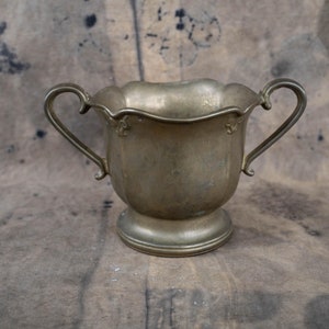 Peut inclure: Un sucrier vintage de couleur argentée avec deux poignées incurvées et un bord festonné. Le bol repose sur une base ronde. L'arrière-plan est de couleur neutre et mouchetée.