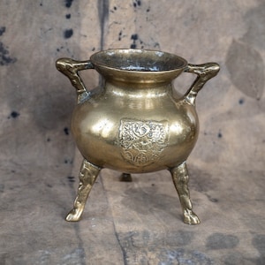 Può includere: Un piccolo vaso antico in ottone con tre gambe e due manici. Il vaso presenta un design ornamentale con uno stemma sul davanti.