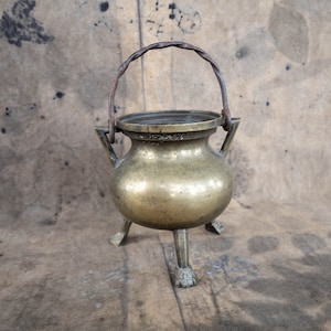 Può includere: Un piccolo calderone antico in ottone con tre gambe e un manico in metallo intrecciato. Il calderone ha un corpo arrotondato e un'ampia apertura. L'ottone ha una calda tonalità dorata e il manico è marrone scuro.