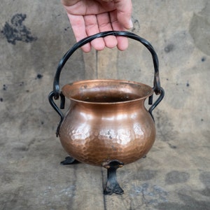 Może przedstawiać: Miedziany kociołek z młotkowaną powierzchnią, z czarnym kutym żelaznym uchwytem i trzema dekoracyjnymi nóżkami. Miedziany garnek ma zaokrąglony korpus i teksturowaną powierzchnię. Uchwyt jest łukowaty i przymocowany za pomocą zakrzywionych podpór. Rustykalny przedmiot w stylu vintage.