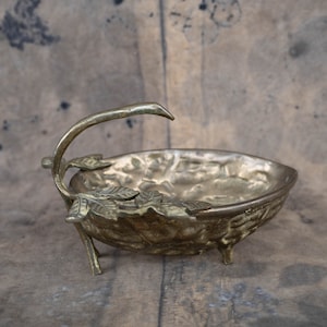 Può includere: Una ciotola decorativa vintage color ottone con un design a foglie. La ciotola ha un manico curvo ed è sostenuta da tre piccole gambe. L'interno della ciotola ha una superficie strutturata. Il design generale ricorda la natura.