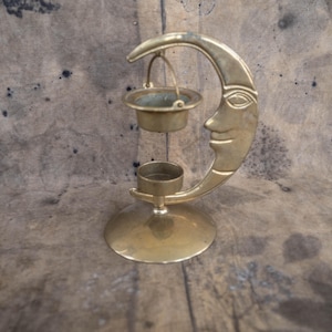 Könnte beinhalten: Ein goldfarbener Metall-Räucherofen in Form eines Halbmondes mit einem Gesicht. Der Räucherofen hat eine kleine hängende Schale für Räucherstäbchen und einen Kerzenhalter auf der Basis.