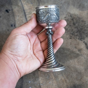 Könnte beinhalten: Ein silberner Kelch mit einem verzierten Design und einem gedrehten Stiel. Der Kelch wird in einer Hand gehalten.