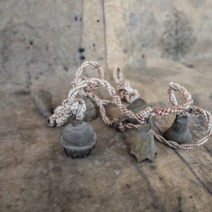 May include: A collection of small, weathered gray bells with a twisted rope. The bells have a rustic appearance, with a variety of shapes and sizes. The rope is white with red accents, adding a touch of color to the scene.