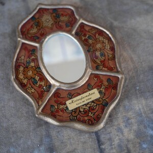 An oval-shaped, wall mirror with a silver-colored frame. The mirror's border features a floral design in red, gold, and blue. A small label on the mirror reads "Hinterglasmalerei Kunst der Völker."