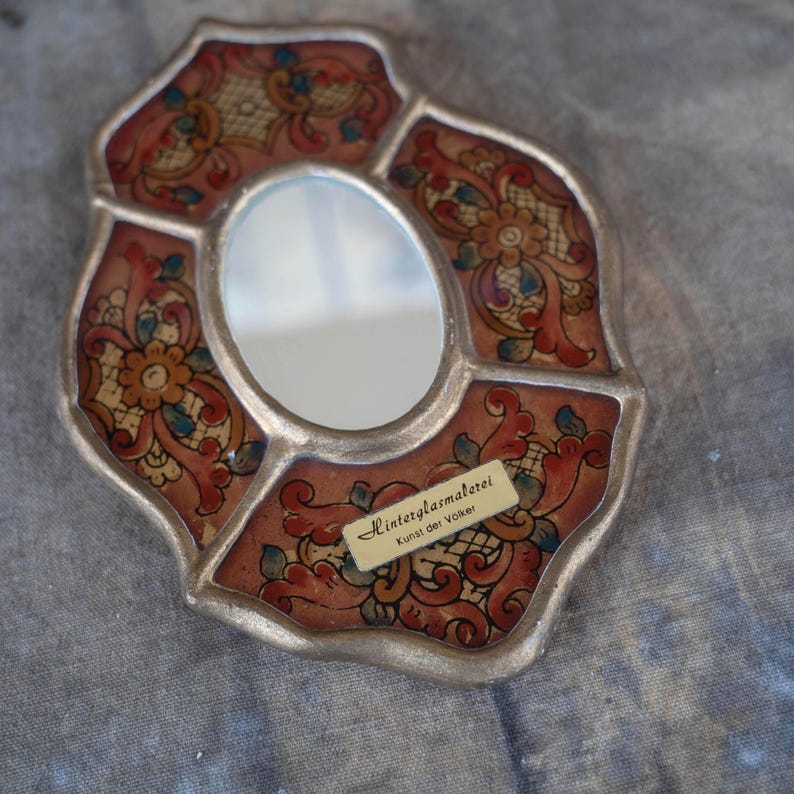 An oval-shaped, wall mirror with a silver-colored frame. The mirror's border features a floral design in red, gold, and blue. A small label on the mirror reads "Hinterglasmalerei Kunst der Völker."