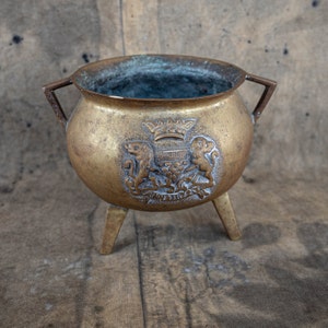 Può includere: Un pentolino in ottone con tre gambe e uno stemma decorativo con una corona e due leoni. Il pentolino ha una patina scura e si trova su una superficie di legno.